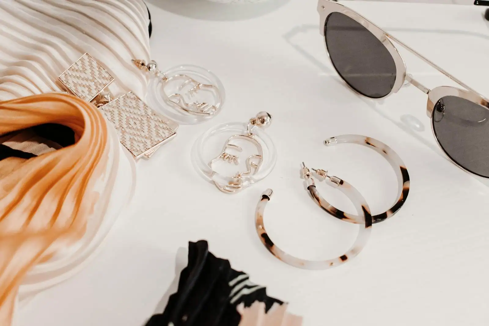 Sélection d'accessoires posés sur une table claire : sac structuré en cuir camel, paire de créoles dorées, foulard imprimé soie, lunettes de soleil