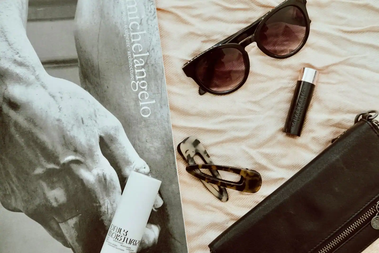 Lunettes de soleil, pochette en cuir, barrette et cosmétiques disposés à plat près d'un livre d'art