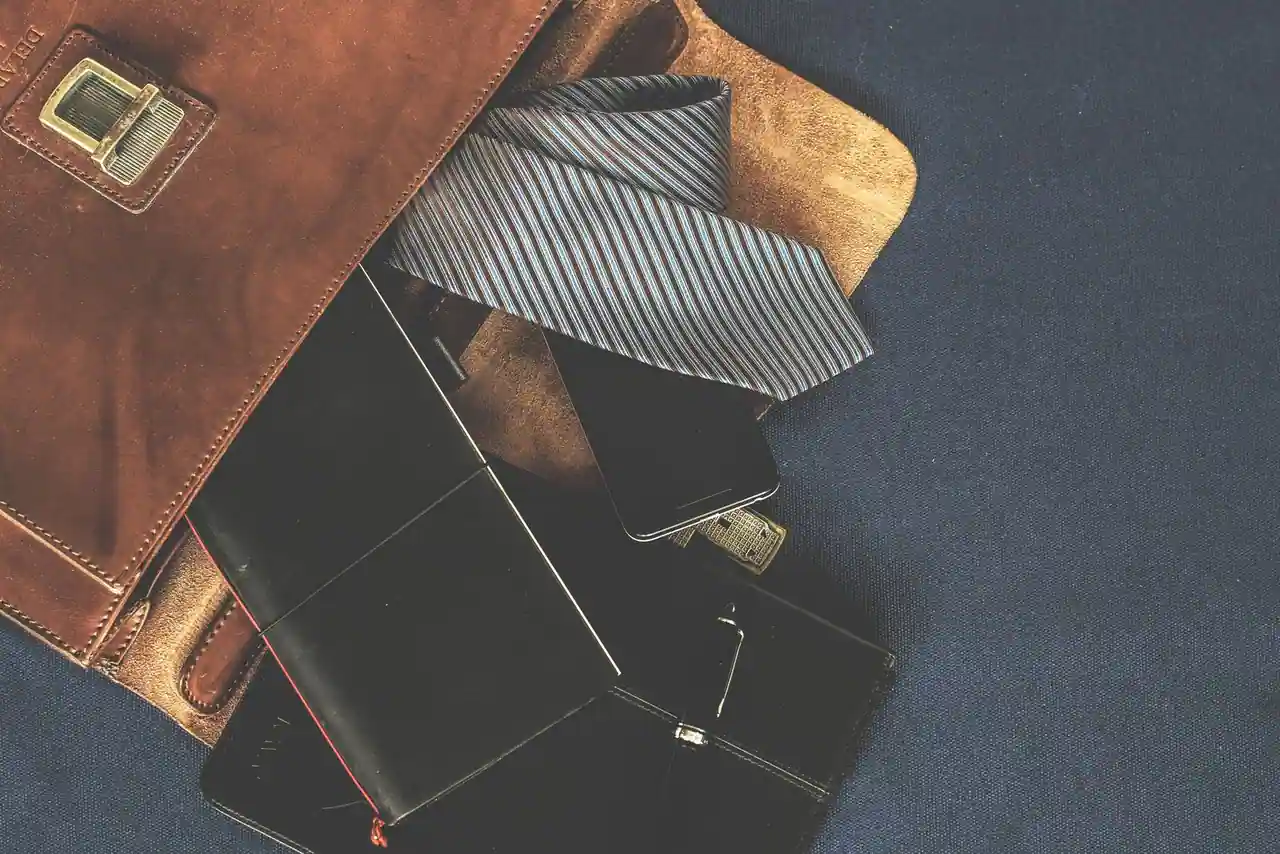 Accessoires masculins disposés à plat : sacoche en cuir, cravate rayée, portefeuille noir et stylo.