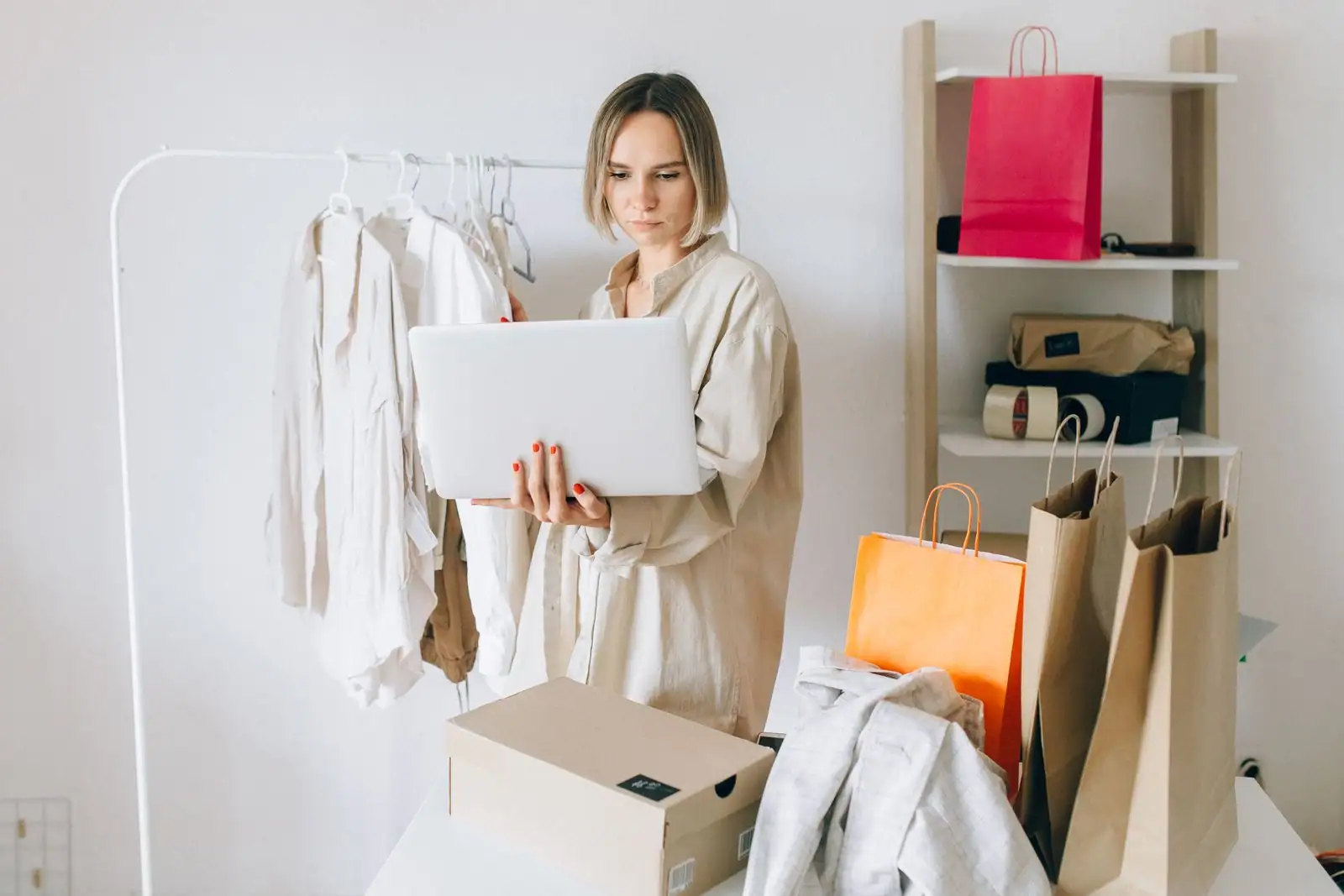 Femme tenant un ordinateur portable près d'un portant de vêtements, avec des sacs et un carton de livraison