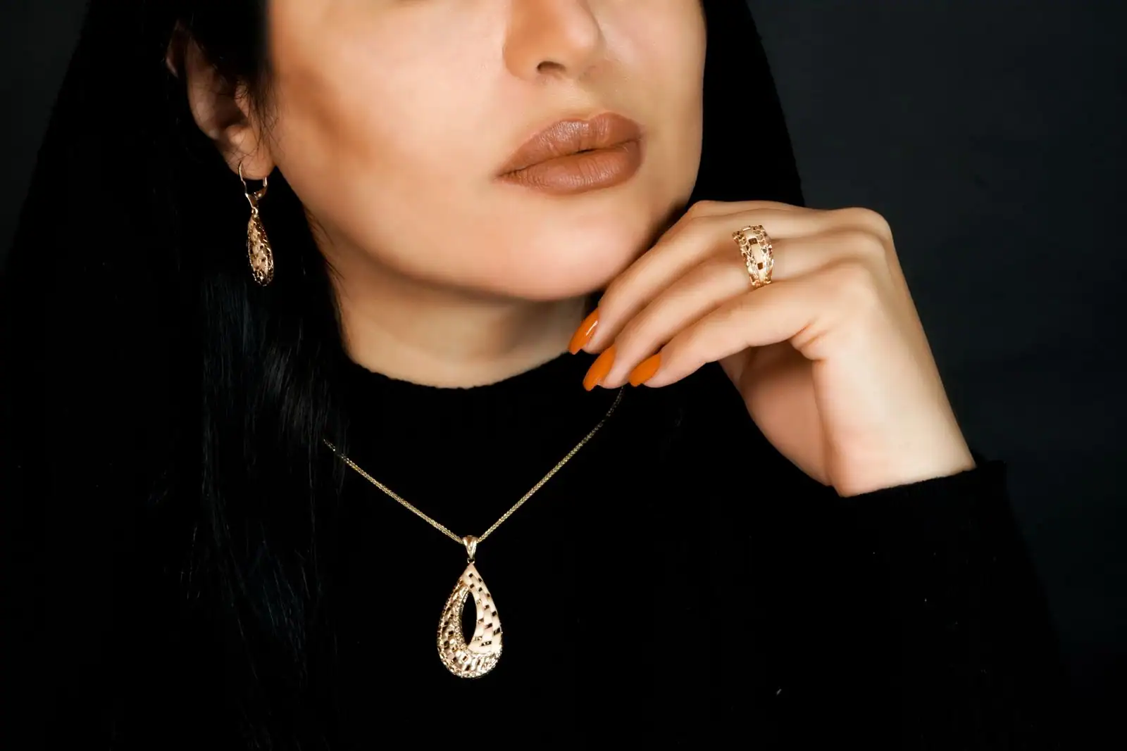 Femme en noir portant une parure dorée assortie : collier à pendentif, boucles d'oreilles et bague