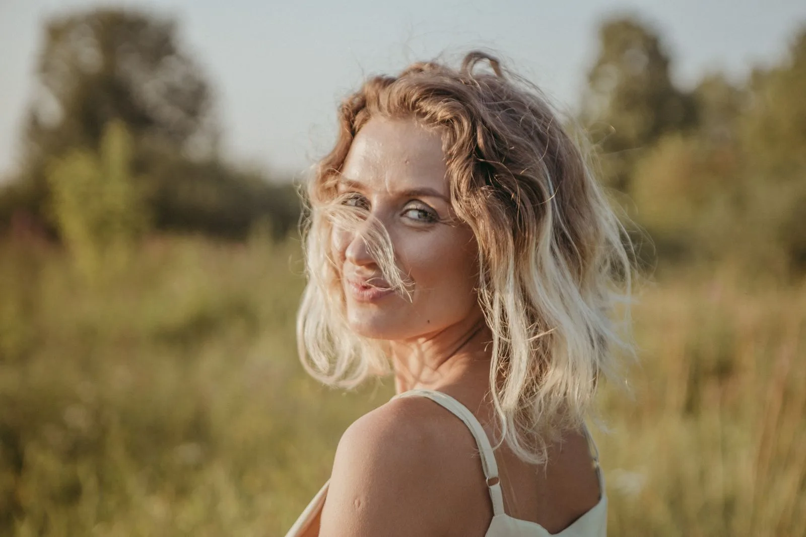 Portrait d'une femme aux cheveux mi-longs blonds et ondulés, tournée vers l'objectif dans un champ ensoleillé