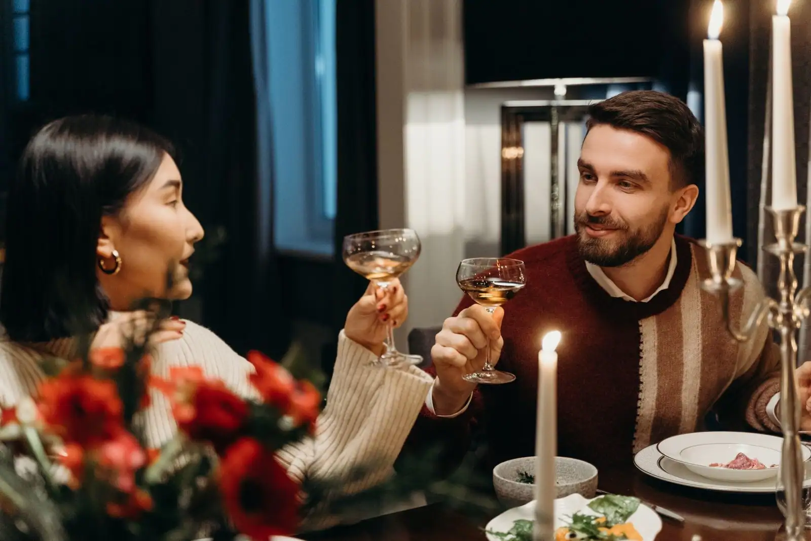 Un couple trinque, deux verres levés, lors d'un dîner aux chandelles dans un cadre intime et chaleureux.
