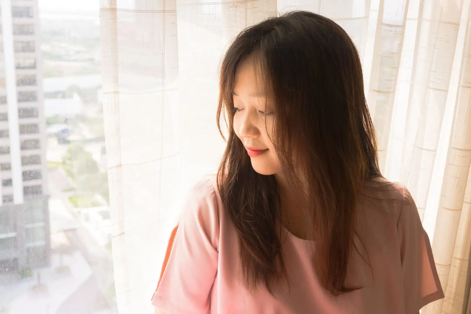 Femme sereine en chemisier rose, debout près d'une fenêtre dans une lumière douce, le regard apaisé
