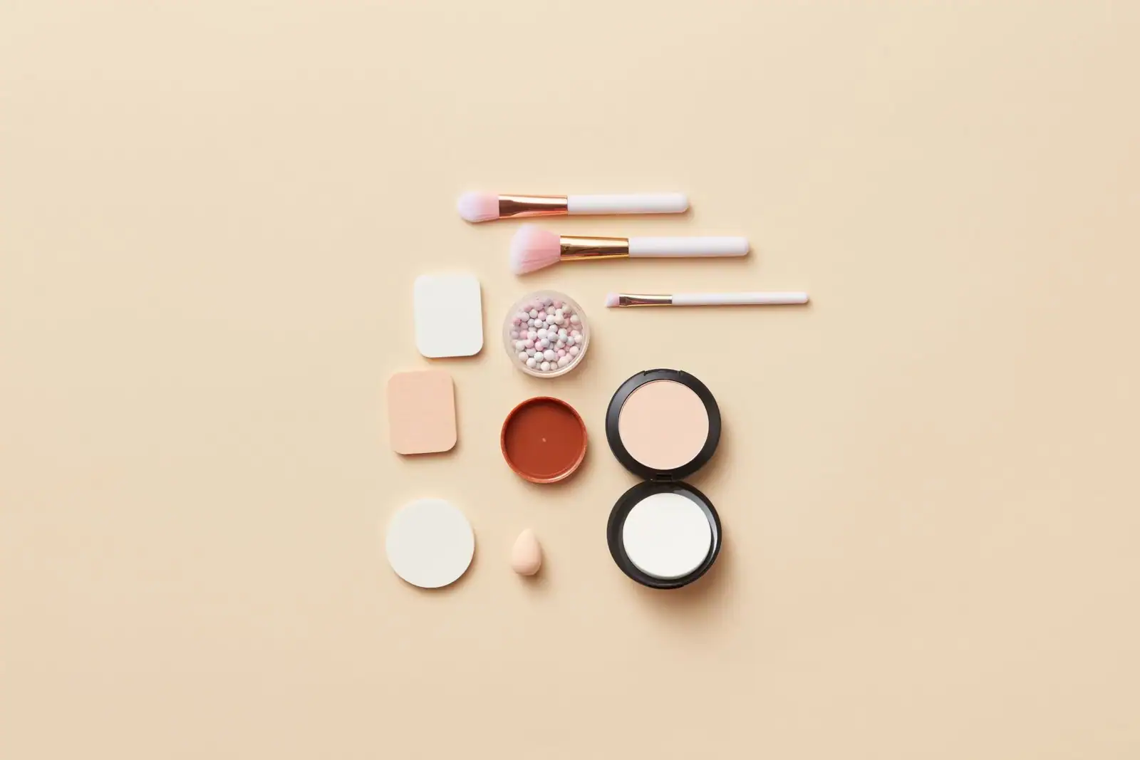 Pinceaux, éponges et poudre compacte de maquillage disposés à plat sur fond beige