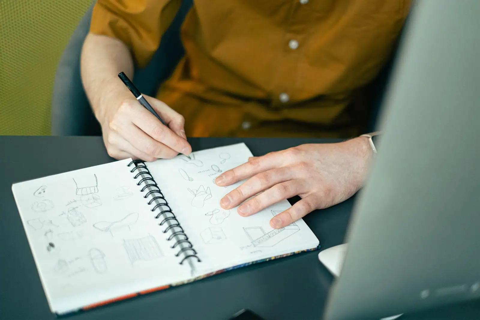 Une personne dessine au stylo de petits croquis dans un carnet à spirale posé sur un bureau, à côté d'un écran