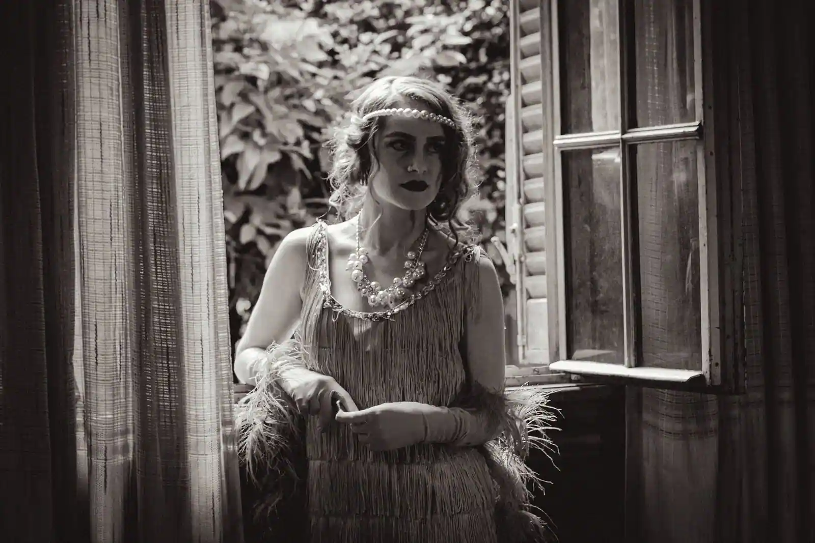 Femme en tenue flapper des années 20 près d'une fenêtre : bandeau perlé, yeux charbonneux et lèvres sombres.