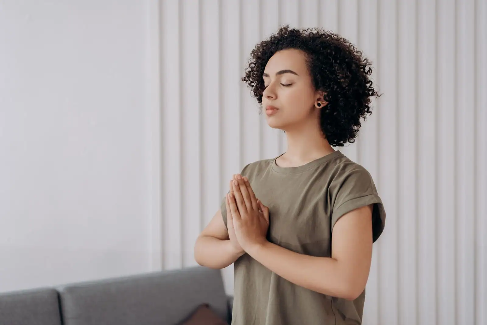 Jeune femme aux yeux fermés et aux mains jointes, dans un moment de méditation au calme chez elle.