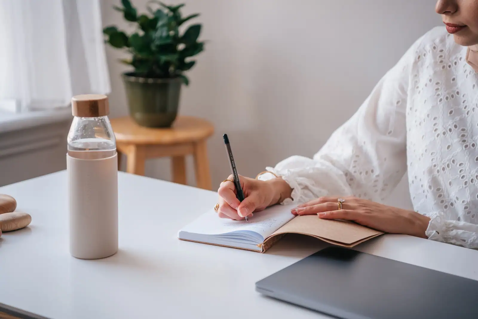 Femme en train d'écrire dans un carnet à un bureau clair, stylo en main, gourde et plante à proximité