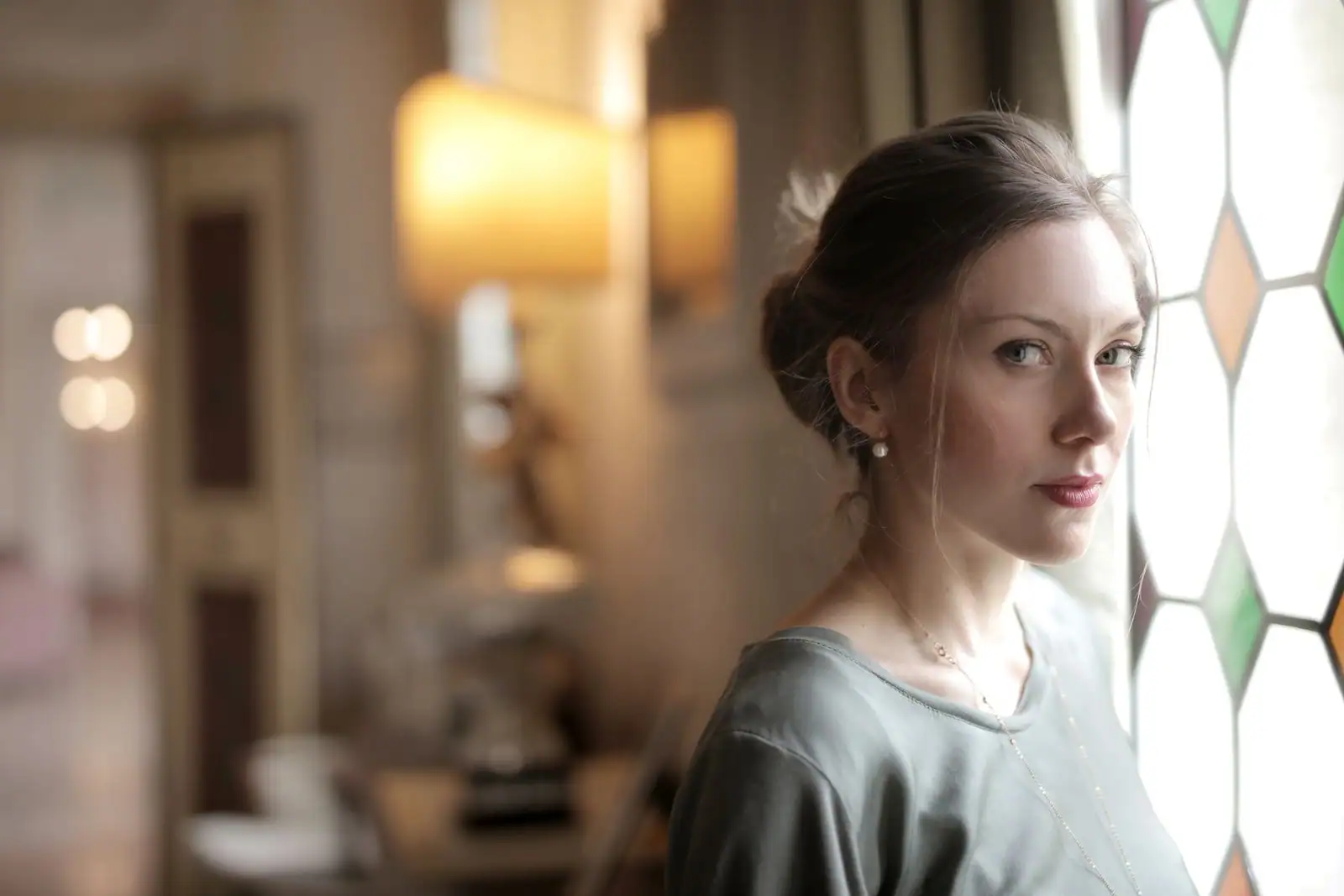 Une femme au visage pensif près d'une fenêtre à vitraux, dans une lumière douce d'intérieur