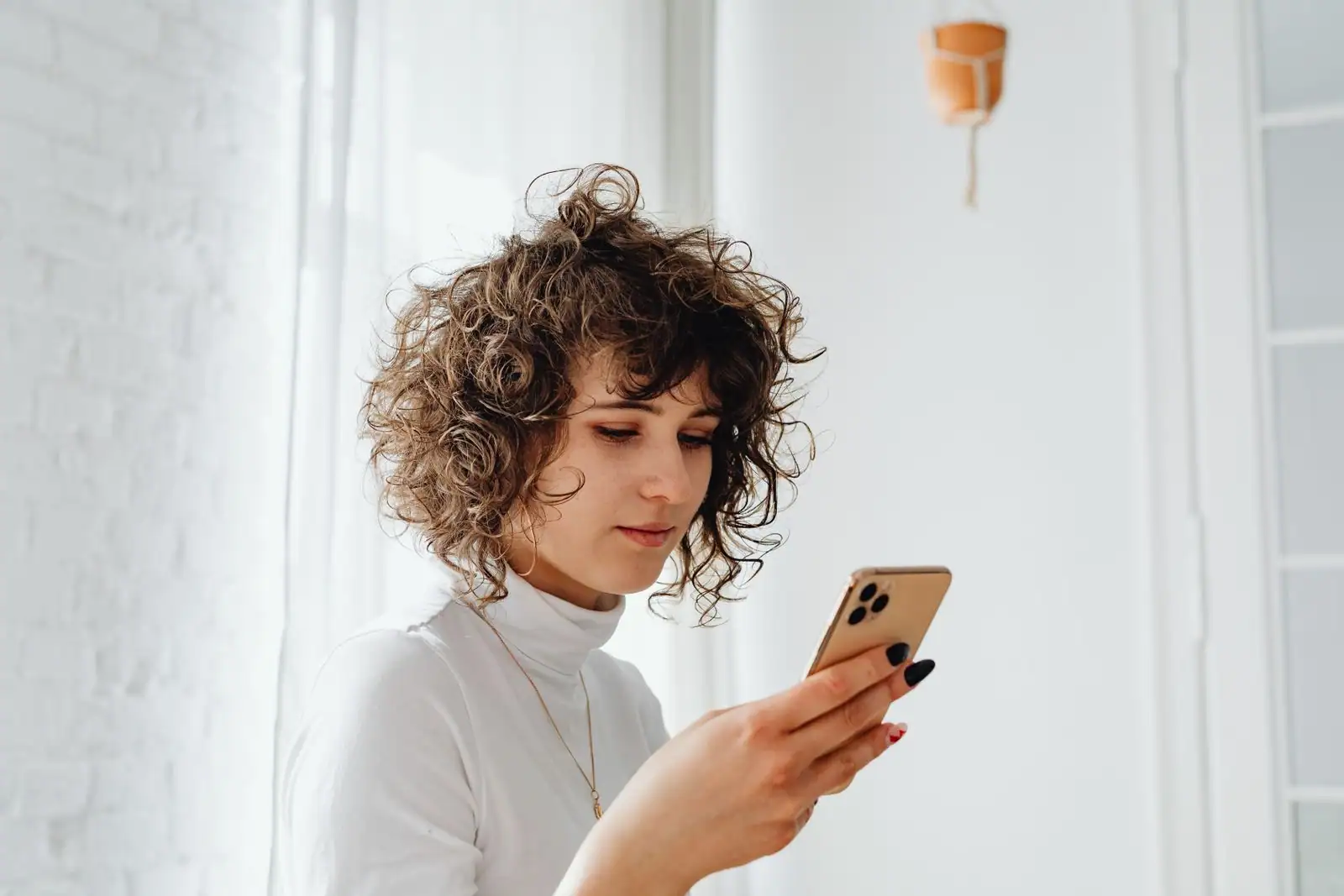 Une jeune femme consulte son smartphone près d'une fenêtre, dans un intérieur clair, l'air concentré.