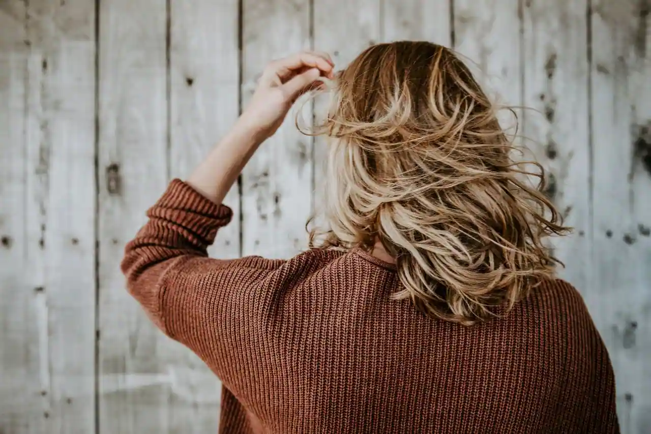 Femme de dos aux cheveux ondulés blond foncé aux reflets clairs, main dans les cheveux devant un mur en bois.