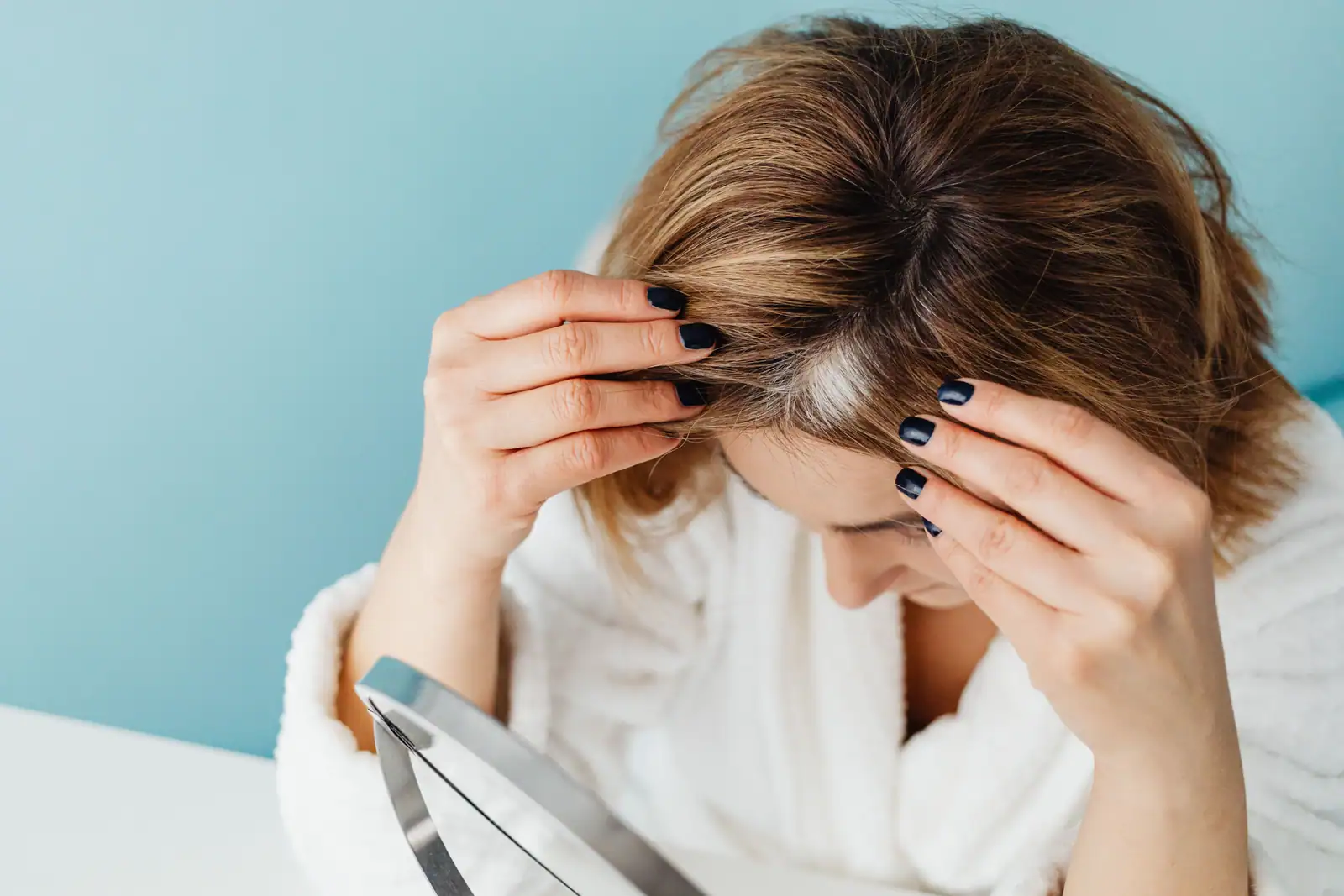Une femme écarte sa raie devant un miroir, laissant voir les cheveux gris apparus à la racine.