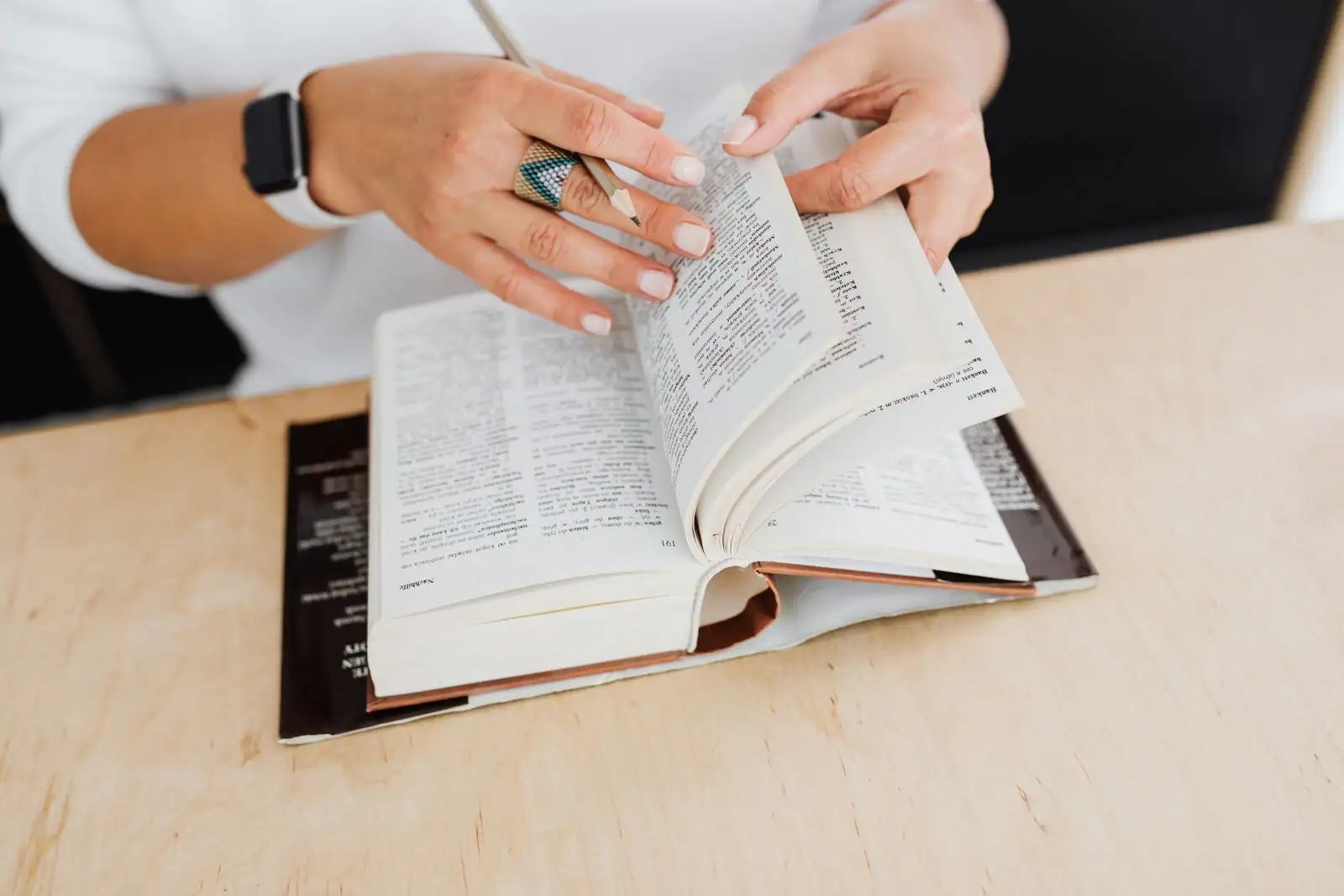 Mains feuilletant les pages d'un dictionnaire ouvert sur une table claire, un crayon entre les doigts.