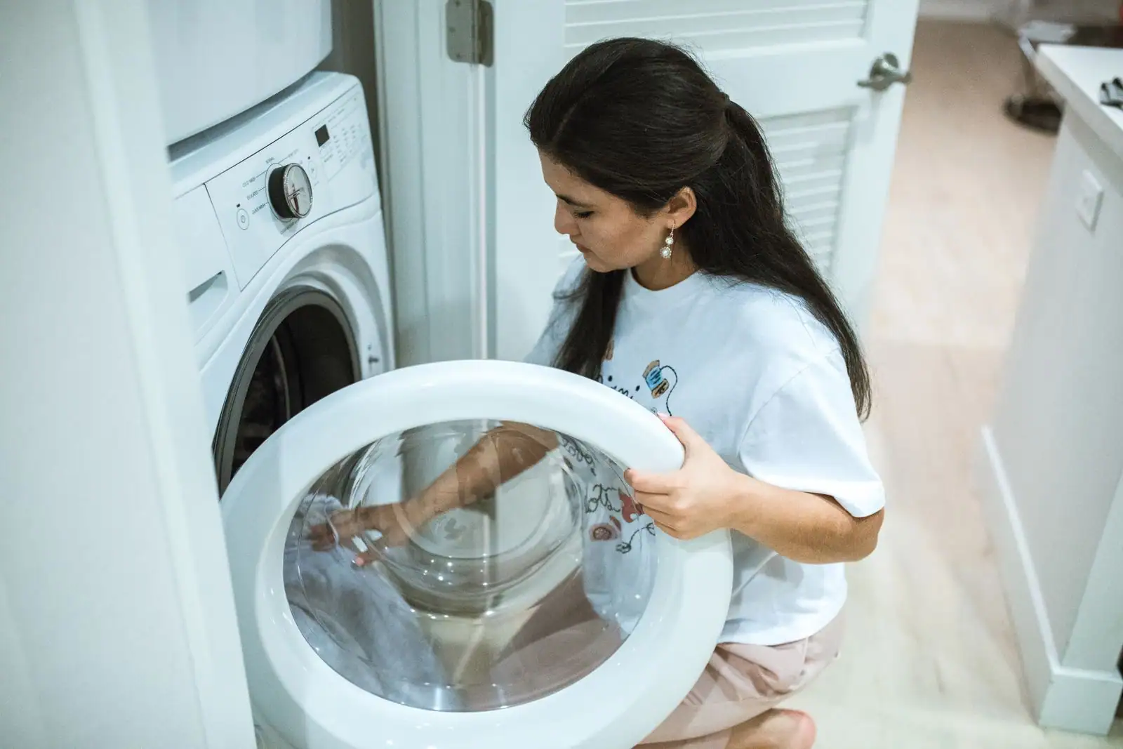 Femme accroupie ouvrant la porte d’une machine à laver pour y charger du linge, à la maison.