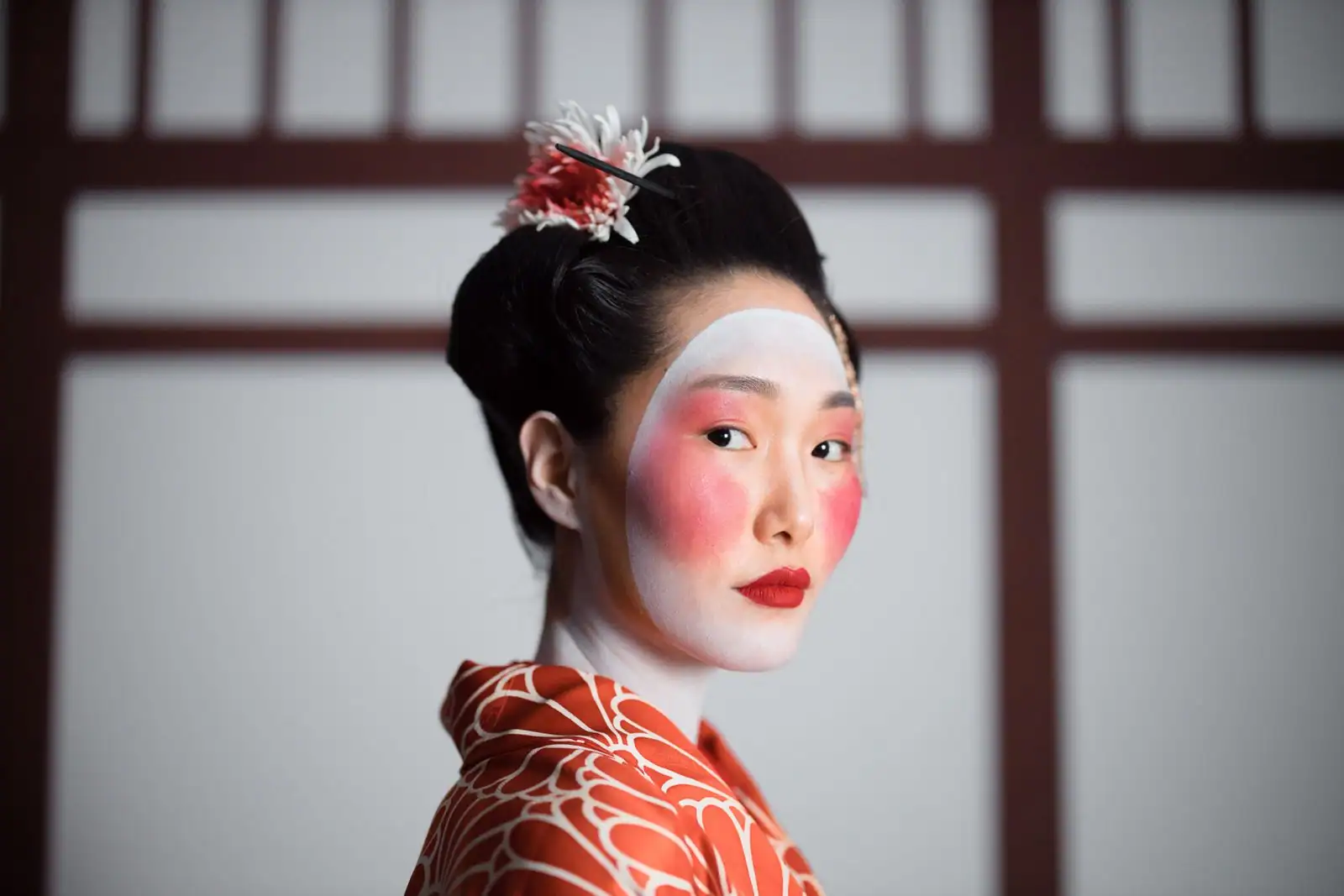 Femme au visage poudré de blanc, joues rosées et bouche rouge, en kimono orange devant un panneau japonais.