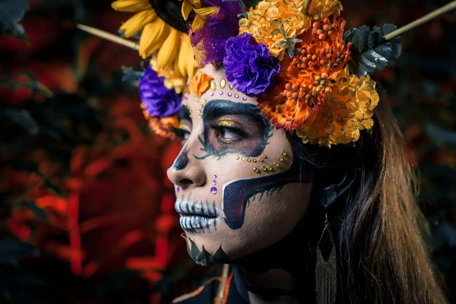 Femme au maquillage coloré de tête de mort mexicaine, coiffée d'une couronne de fleurs orange et violettes.