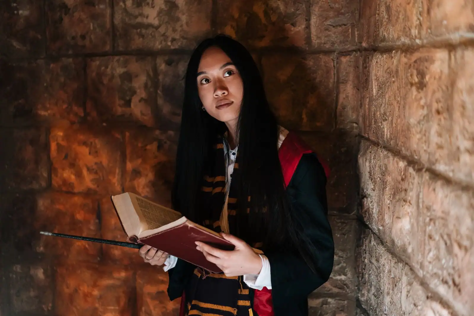 Jeune femme costumée en élève sorcier, écharpe rayée rouge et or et grimoire en main, devant un mur de château.