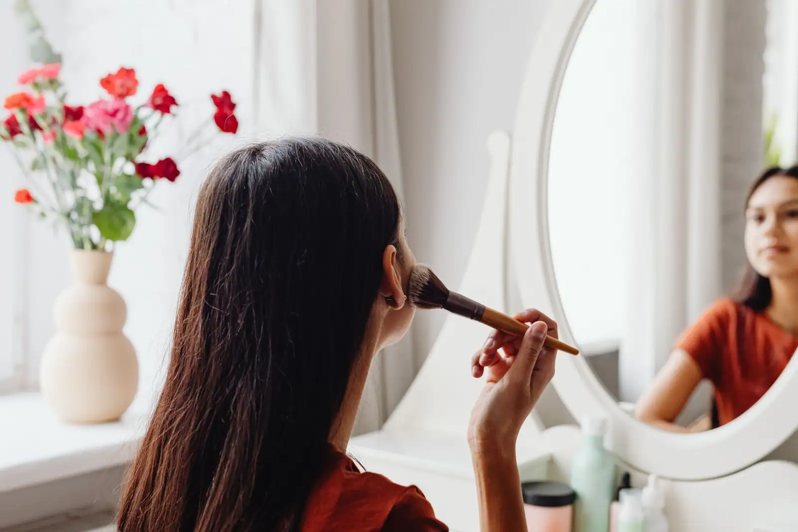 Femme brune appliquant du maquillage au pinceau devant le miroir d'une coiffeuse, un bouquet de fleurs posé à côté.