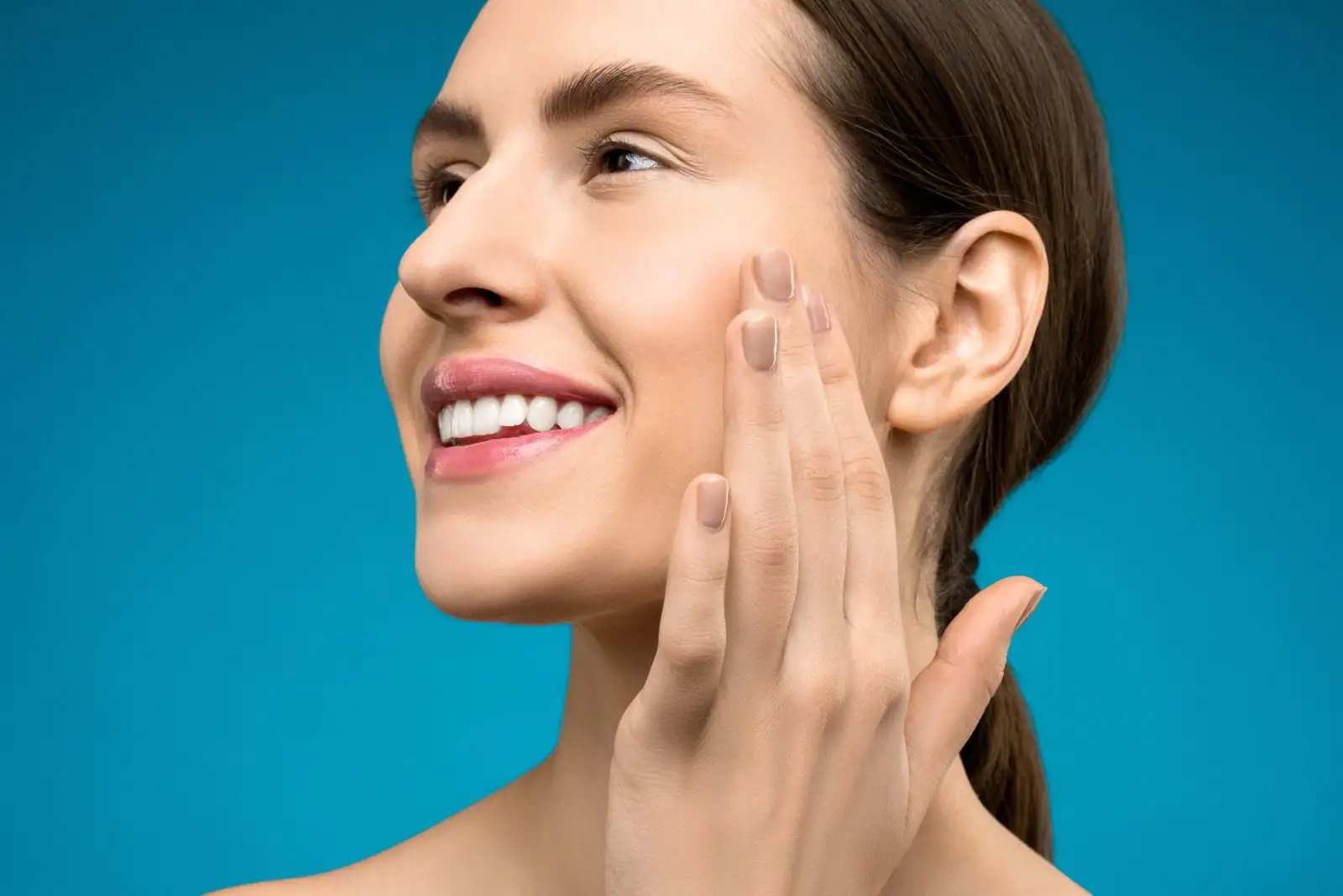 Femme souriante de profil au teint lumineux et au maquillage naturel, la main posée sur la joue.