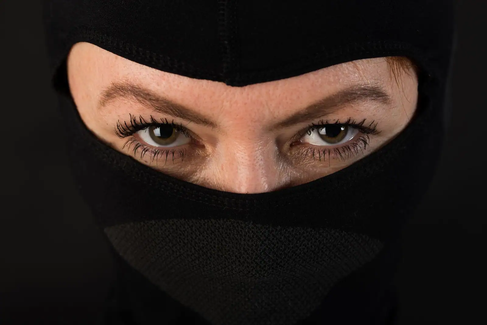 Visage masqué d'une cagoule noire de ninja, seuls les yeux maquillés au mascara sont visibles, en gros plan.