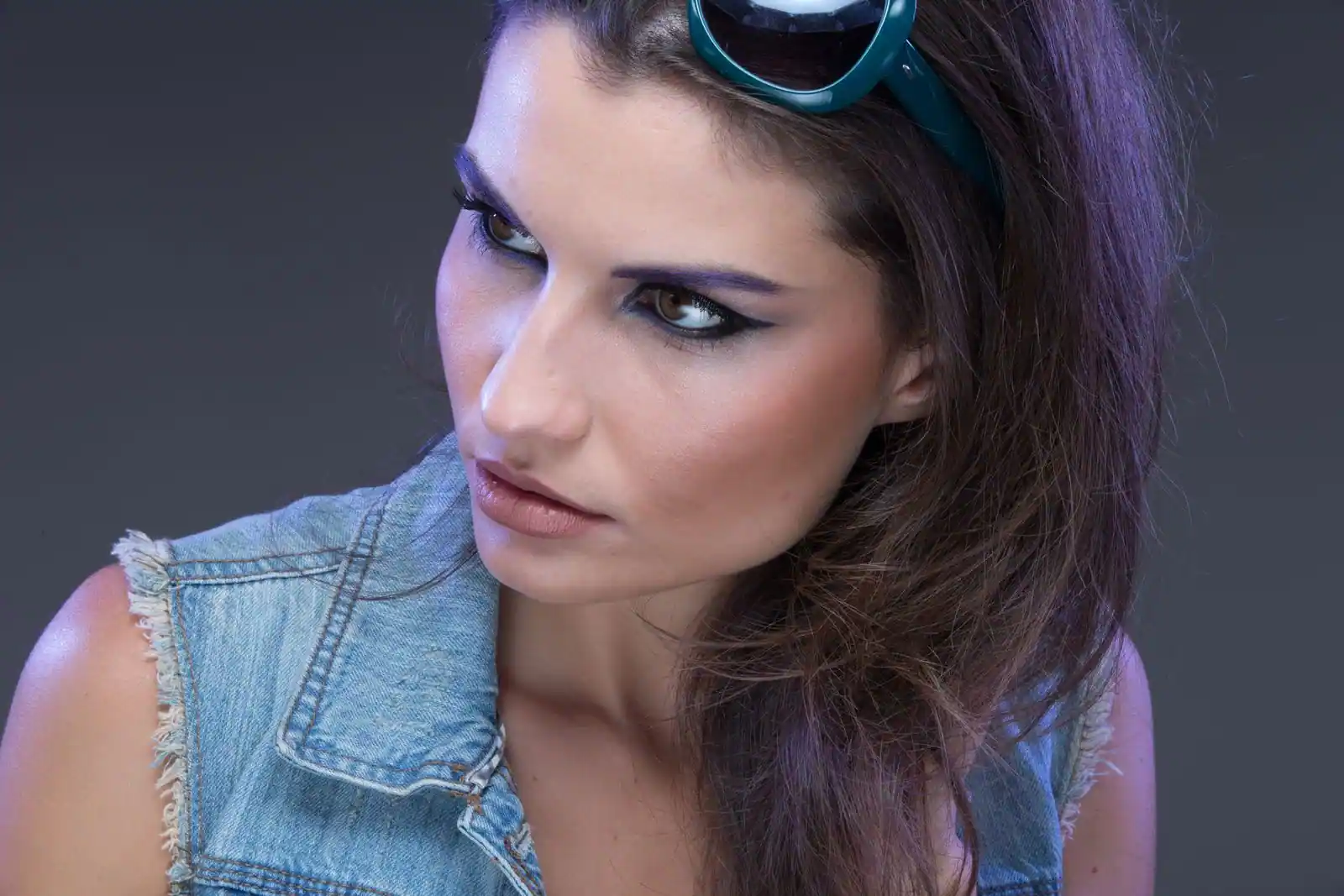 Jeune femme au regard charbonneux marqué, veste en jean et lunettes de soleil relevées.