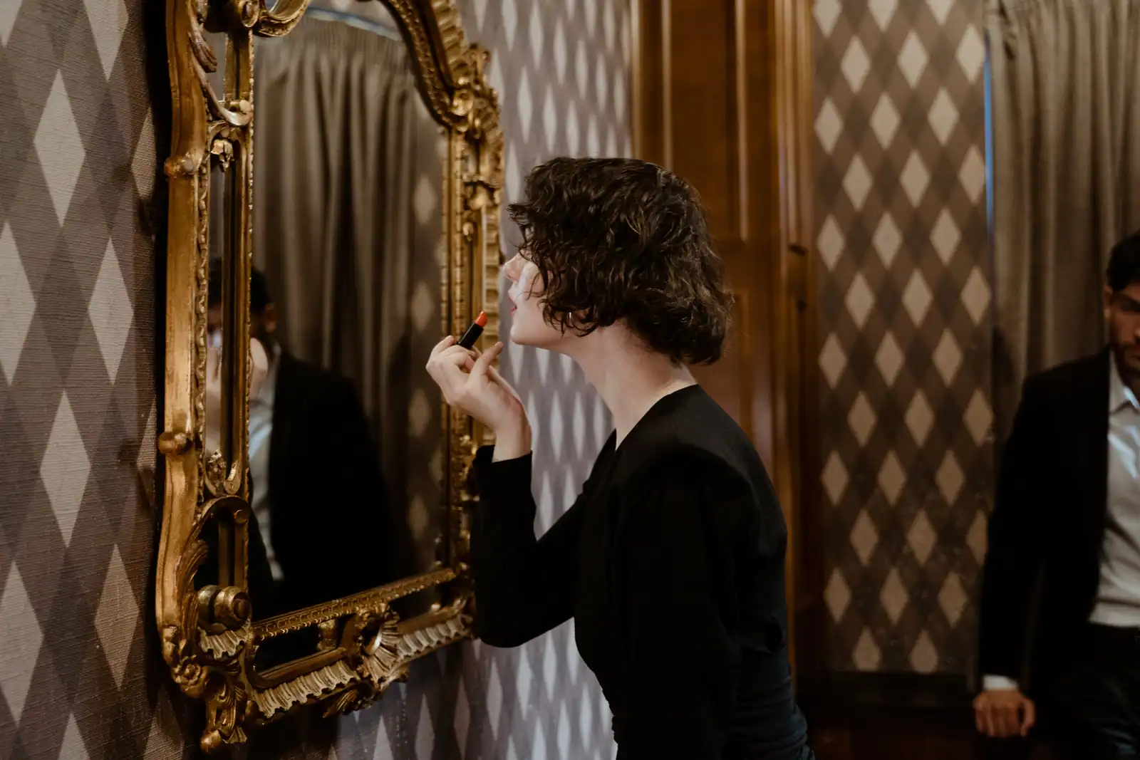 Femme en noir applique un rouge à lèvres devant un miroir doré, la bouche comme unique point fort du visage.