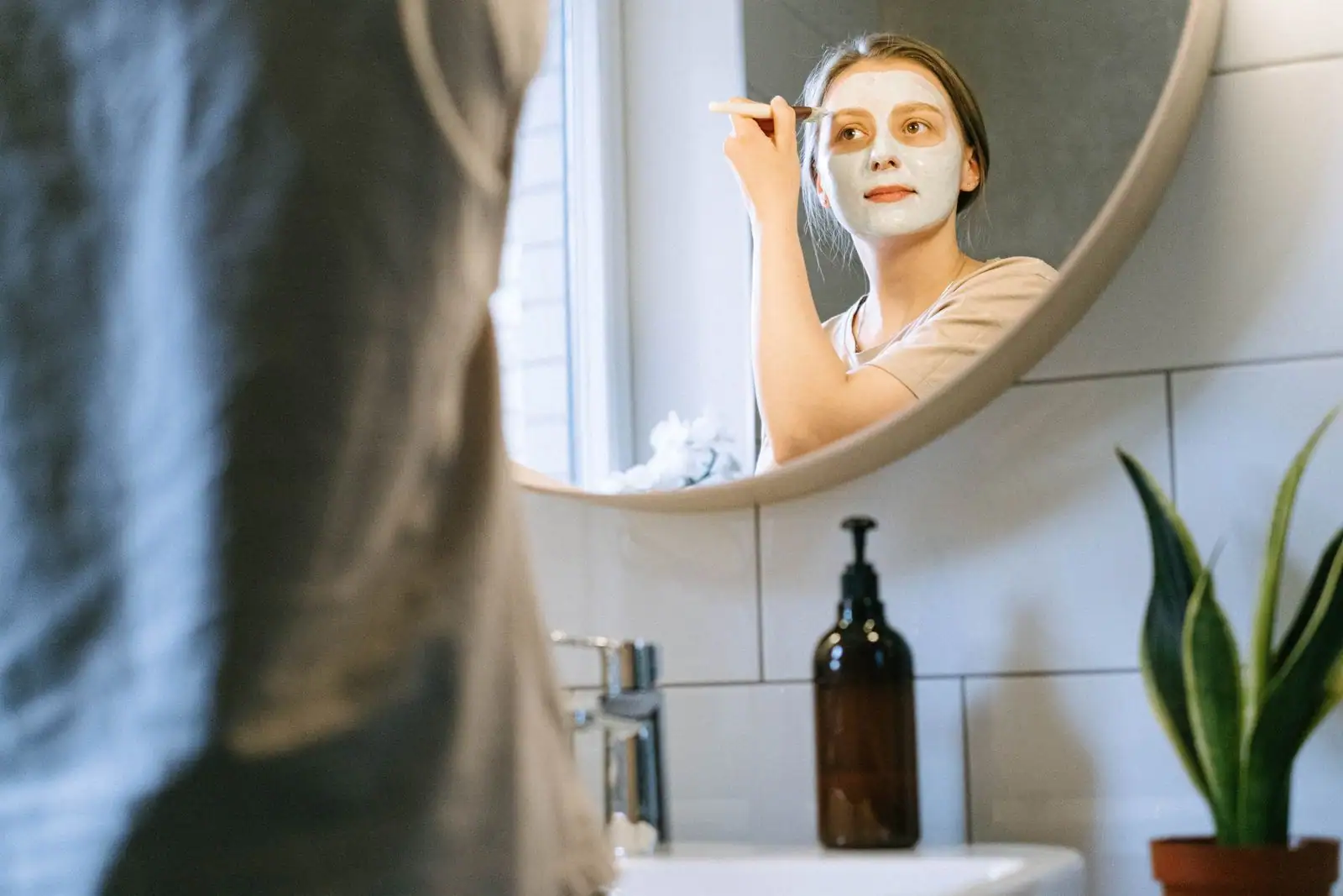 Femme appliquant un masque à l'argile sur son visage au pinceau, devant le miroir d'une salle de bains.