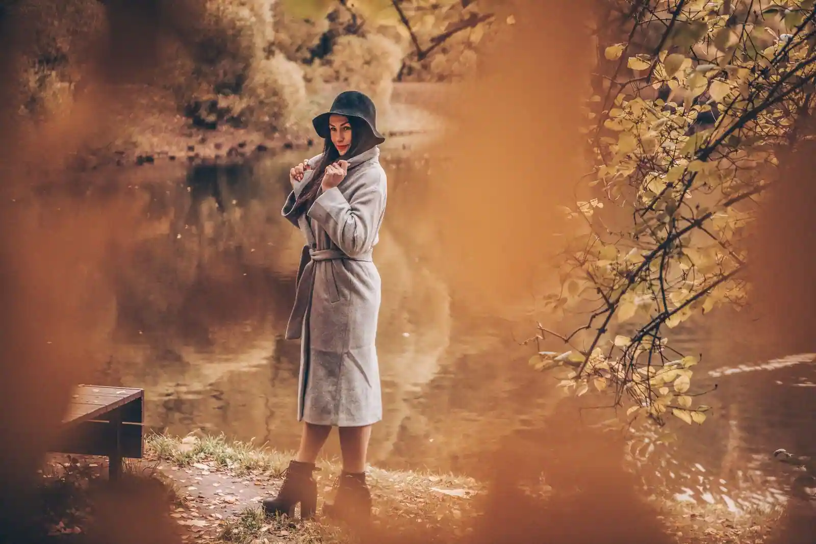Femme en long manteau de laine gris et chapeau, bottines aux pieds, au bord d'un étang aux tons d'automne.