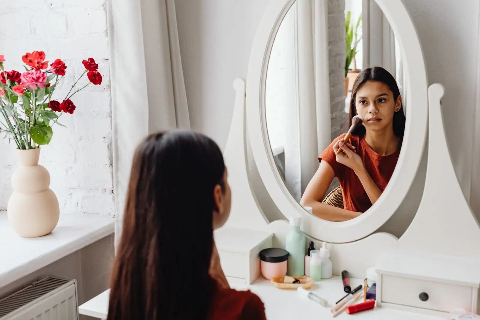Jeune femme appliquant son maquillage au pinceau devant le miroir d'une coiffeuse, cosmétiques posés sur la tablette.